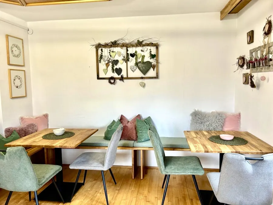 Das Café bei hoamelig: Holztische, grüne Polsterstühle und eine Wandbank mit Kissen, an der Wand ein Rahmen mit Herzen.