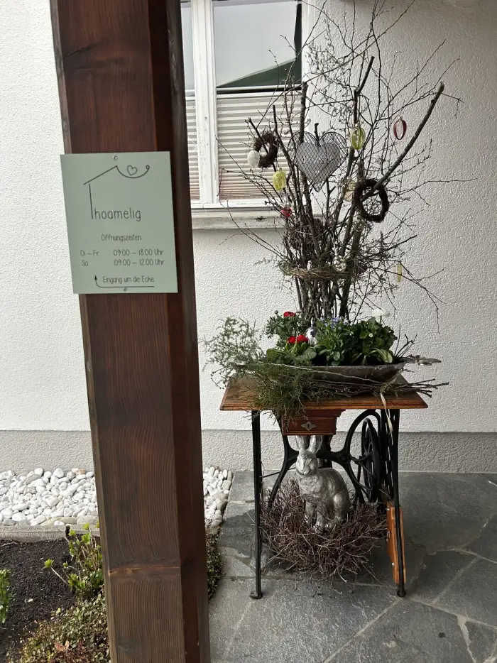Der Eingang zu hoamelig: Schild am Holzpfosten, daneben ein Nähmaschinentisch mit Frühlingsdeko und Metallhase.